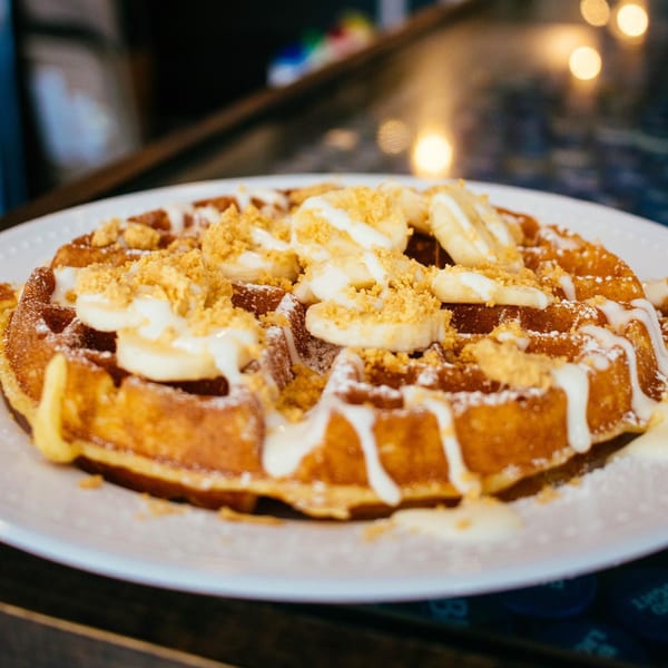 Golden waffle topped with banana slices, white icing drizzle, and golden crumble on a white plate.