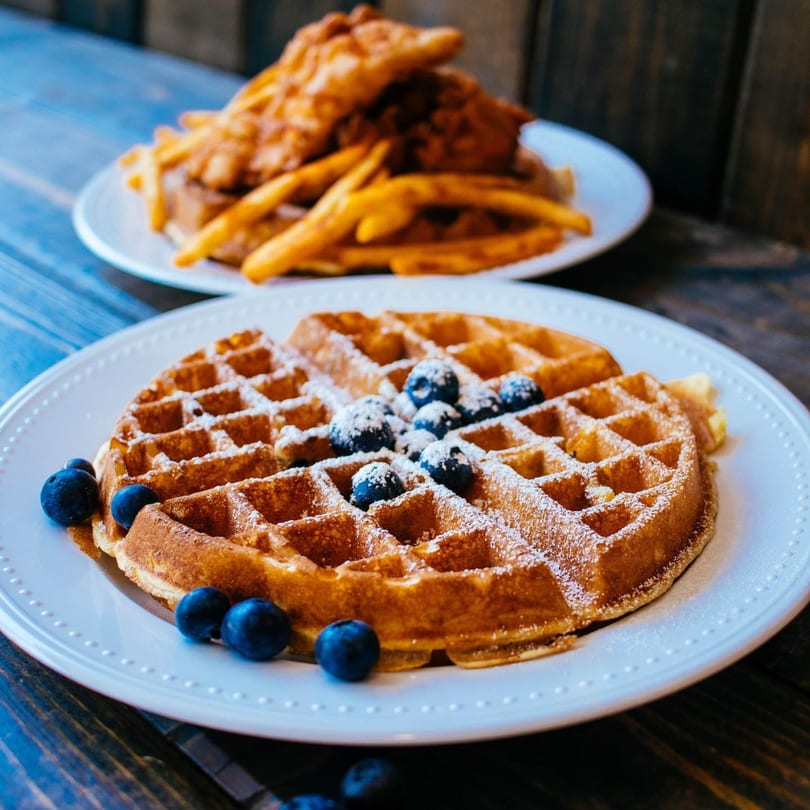 A waffle with blueberries and powdered sugar in the foreground, with chicken and fries blurred in the background.
