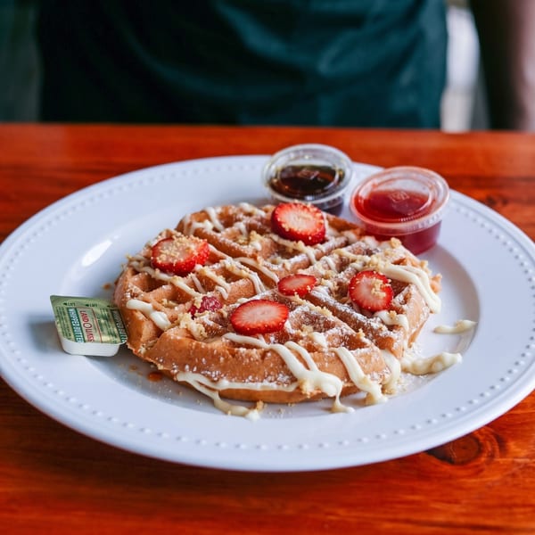 Waffle topped with strawberries, white icing, powdered sugar, and two small cups of syrup.