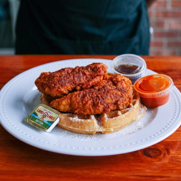 Crispy fried chicken tenders on a waffle with powdered sugar, butter, syrup, and hot sauce.